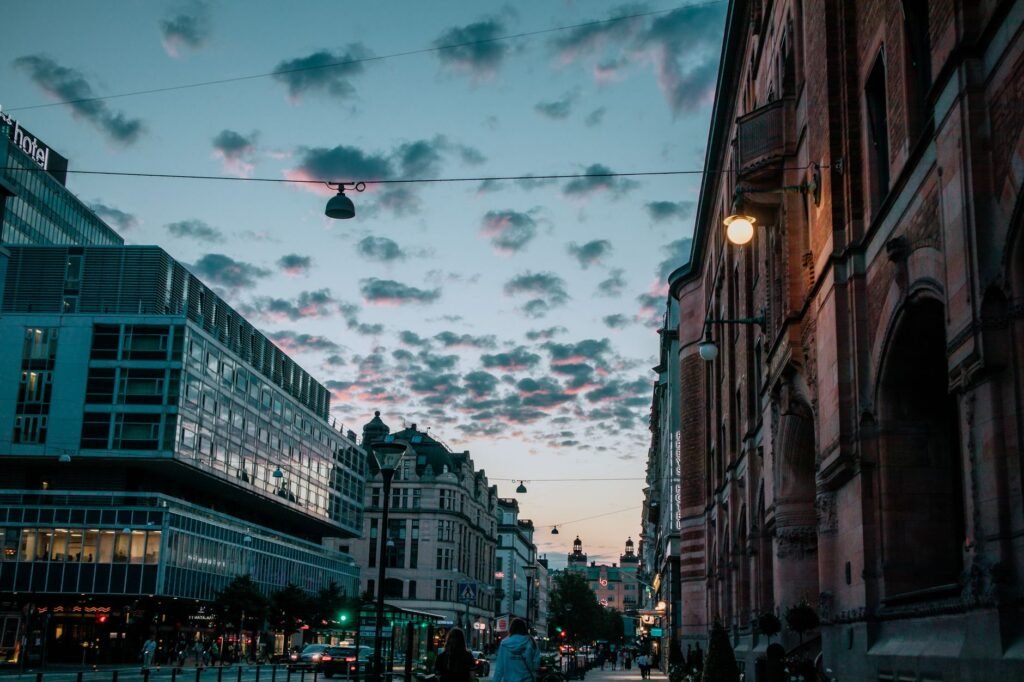 A city street at dusk with warm lights and pedestrians heading to evening venues